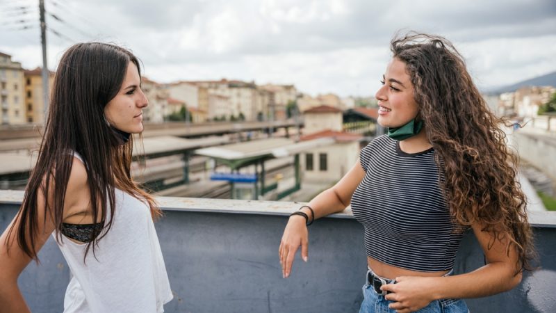 Two women talking on a roof top, one with a mask pulled below her face